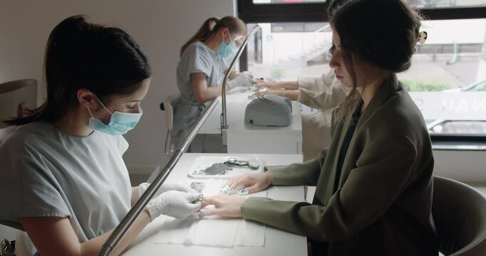 Nail studio with professional masters doing manicure for female clients. Manicurist in mask and gloves treats cuticle with pusher at beauty salon. Hand care procedures. Manicure in a beauty salon.