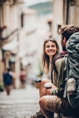 Fototapeta premium Lovely couple romantic moment. A man and woman sitting on a cobblestone street, both wearing backpacks and holding coffee cups. The background is slightly blurred, revealing a few people, buildings.