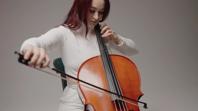 A beautiful woman sitting and playing the cello in a studio on a white background. Classical music playing stringed classical musical instruments, Female cellist practicing solo piece.