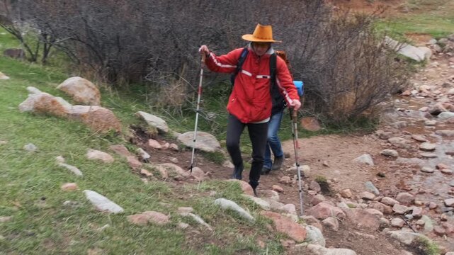 hikers walking on rocky trail front view, two men trekking uphill after stream crossing using poles and carrying backpacks, regaining balance on uneven ground, outdoor endurance and terrain navigation