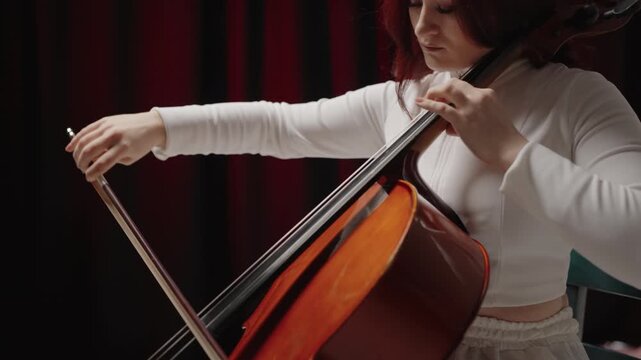 A beautiful woman sitting and playing the cello in a studio on a black background. Classical music playing stringed classical musical instruments, Closeup view of violoncello and hands of musician,