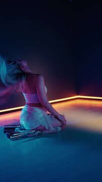 Sensual blonde woman in white outfit performing a dynamic and flexible floorwork contemporary dance routine in a dark studio with blue and orange neon lights, moving gracefully in slow motion.Vertical