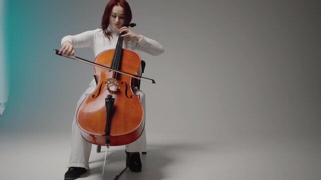 A beautiful woman sitting and playing the cello in a studio on a white background. Classical music playing stringed classical musical instruments, Female cellist practicing solo piece.