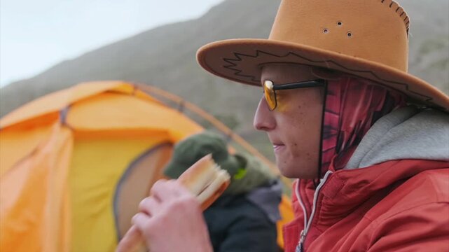 hikers eating food during rest near tent, outdoor camping scene in mountains, close lifestyle moment with natural environment, travel activity and relaxation after hiking