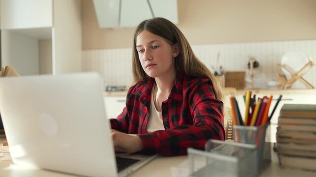 teen studies at desk with laptop girl concentrates on homework and notebook while student writes with pen near pencil cup and book stack in kitchen setting focus education concentration and study