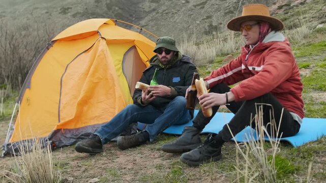 hikers eating sandwiches near tent close-up, two men sitting on mat drinking beer and having meal after hike, relaxing during camping break, outdoor leisure and recovery moment concept