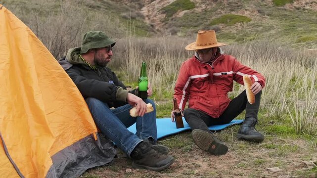 hikers eating sandwiches and drinking beer close-up, two men sitting near tent after hike, resting on mat and sharing simple meal, outdoor relaxation, recovery and companionship moment concept