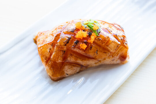 Grilled salmon aburi sushi topped with sweet sauce and tobiko, served on a white ceramic plate over a light wooden table background.