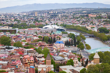 A panoramic view of Tbilisi, Georgia showing the Kura River, the glass Bridge of Peace, concert hall and a dense mix of historic churches and modern buildings across a green urban landscape © evannovostro