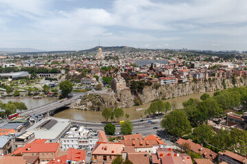 Aerial view of Tbilisi, Georgia showing the Mtkvari river, historic Metekhi cliff, terracotta rooftops and Sameba Cathedral dome amid green trees and urban landscape under a bright cloudy sky © evannovostro