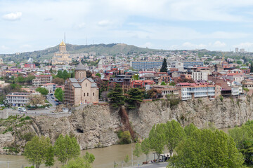 Panoramic view of Tbilisi featuring the historic Metekhi Church on a rocky cliff and the golden-domed Sameba Cathedral in the background, with riverfront trees, colorful houses and urban hillside © evannovostro