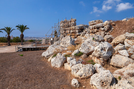 Ggantija ruins, ancient megalithic ruins set in a dry Mediterranean landscape, surrounded by large limestone boulders, palm trees and temporary scaffolding. Malta