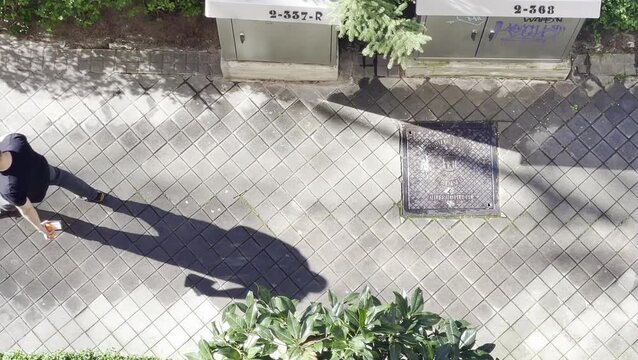 Calvo con tel&eacute;fono m&oacute;vil y caminando con sombra y silueta grande y alargada en acera de calle desde arriba en perspectiva a&eacute;rea