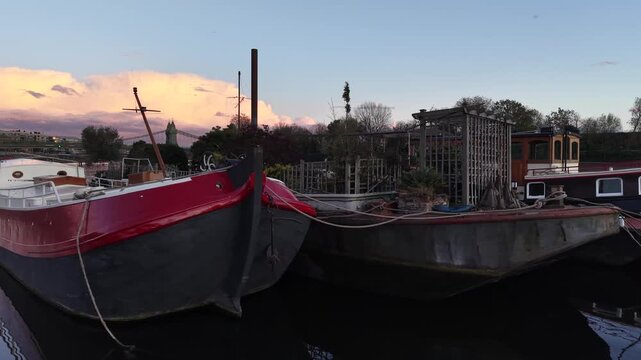 LONDON, UK - JUNE 2026: Video showing classic residential houseboats on the River Thames riverbank during golden hour, revealing Hammersmith Bridge under a dramatic twilight sky