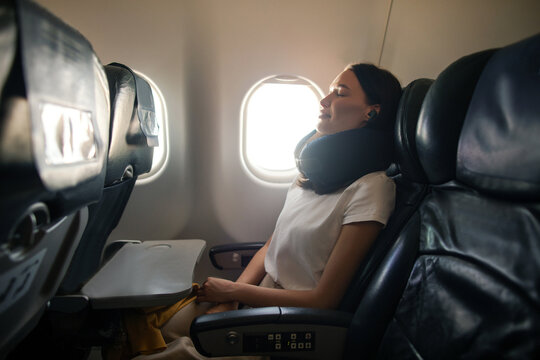 Happy relaxed woman sitting in plane seat with her eyes closed, wearing neck pillow and listening music in earphones, side view shot