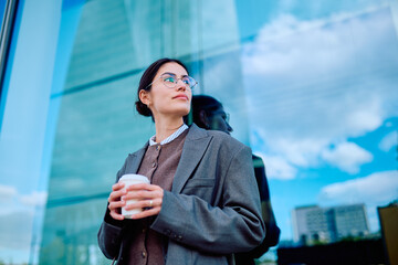 Businesswoman standing outdoors holding a coffee cup, looking thoughtfully towards the urban landscape and reflecting on career goals © we.bond.creations