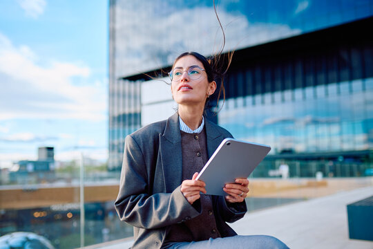 Corporate woman using a digital tablet outdoors, thinking about career growth and ambitions in a modern city environment