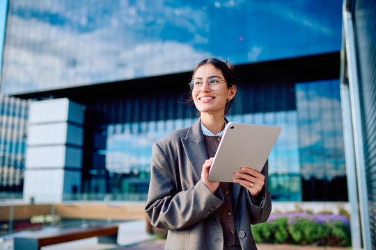 Professional businesswoman smiling, using a digital tablet while standing at a modern office building, symbolizing success and technology