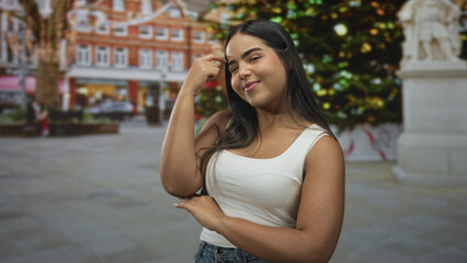 Fototapeta premium Young hispanic woman smiling with hand to cheek in street by decorated christmas tree; confident holiday cheer.