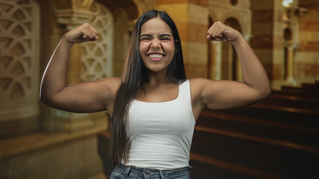 Woman flexes biceps and clenches fists while smiling, wearing white tank top and jeans, in ornate stone building hall with arched columns; confidence empowerment.