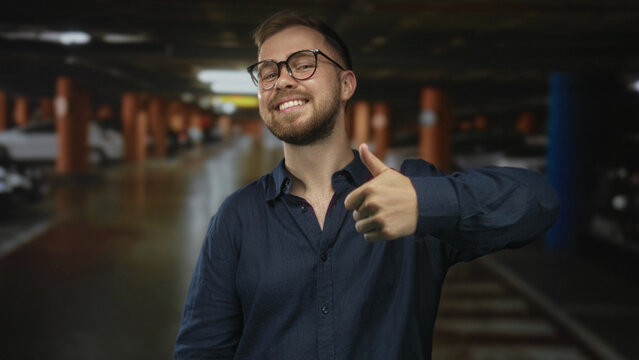 Man with glasses and beard smiling and giving thumbs up inside parking building near cars wearing a navy shirt; optimism.
