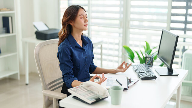 Serene Office Meditation: An Asian woman finds inner peace through meditation at her workspace, balancing work with wellness. Capturing the tranquility amidst daily demands
