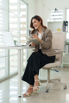 Asian adult professional woman writing notes planning business strategy laptop computer bright corporate office workspace daylight window inspiration productivity focus