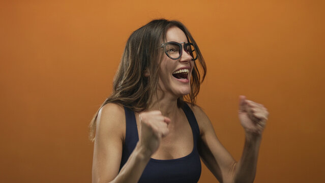 Woman wearing black glasses with clenched fists and a wide grin stands on an orange studio set; excitement.