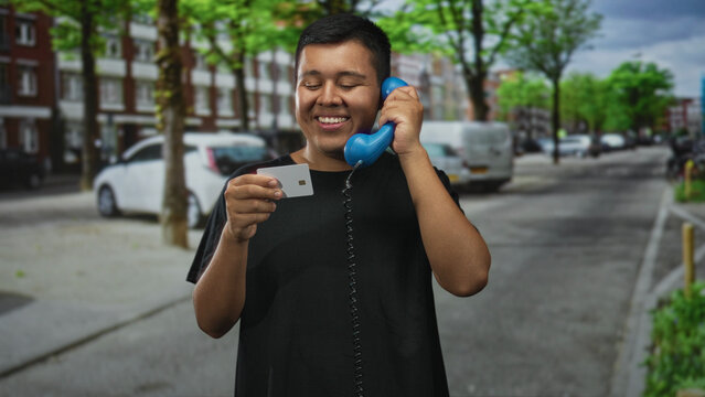 Young man holding a blue handset to his ear and a creditcard on a city street; quiet contentment everyday moment.