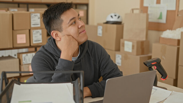 Man smiling resting chin on fists beside laptop and barcode scanner in parcel office building with stacked boxes; contentment entrepreneurship.