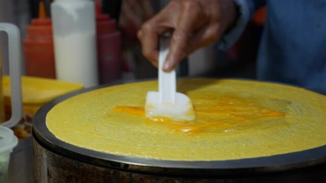 A street vendor spreads savory filling across round pancake batter on a hot cooking surface while preparing Thai style Takoyaki Crispy Pancake at a food stall. Street food.