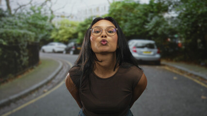 Fototapeta premium Young brunette woman wearing glasses points both index fingers toward camera showing hands and wrists on a leafy residential street; playful confidence.