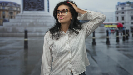 Obraz premium Woman in white shirt and glasses touches hair on wet city street in front of building; contemplation.