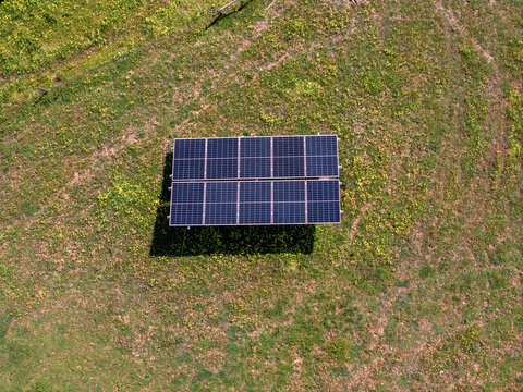 Aerial view of a small solar panel array installed on a green grassy field with scattered yellow wildflowers in a rural area.