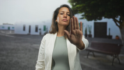 Fototapeta premium Woman holds up hand palm forward for stop in street in front of white building and bench; defiance protection.