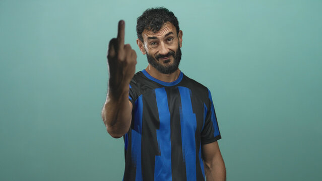 Man in blue and black soccer jersey shows middle finger to camera in studio, bearded player with stern face, extended arm and provocative gesture; defiance protest.