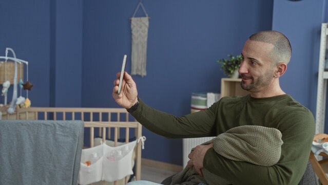 Man holds swaddled baby and raises smartphone for videochat in nursery, smiling while supporting infant against his chest; tenderness bonding.