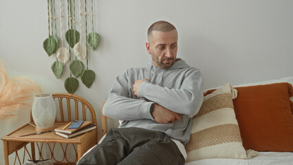 Fototapeta premium Man holds tablet to chest on bed in bedroom, arms crossed and looking down beside rattan side table with books and decorative pillow; quiet contemplation.