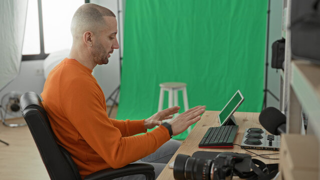 Man photographer pointing at tablet and gesturing with hands at desk in studio, green screen and camera visible; concentration creativity.