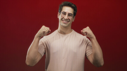 Naklejka premium Man raising clenched fists and smiling in a red studio wearing glasses and a casual t shirt; joy triumph.