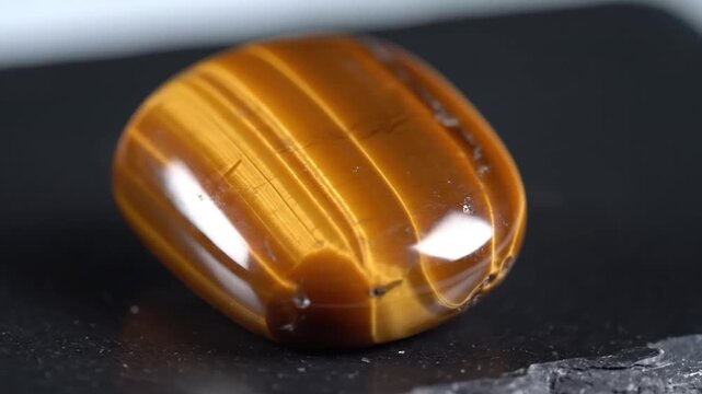Polished tigers eye gemstone showing golden brown chatoyancy stripes, natural mineral with subtle light reflections on dark textured surface