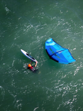 Aerial view of a person in the water wearing an orange helmet and red life vest with wing foiling equipment including a blue wing and board.