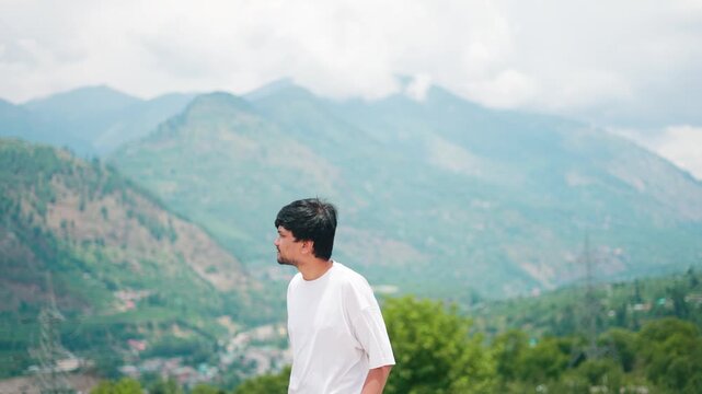 4K Portrait shot of an Indian man walking in front of Himalayan mountain peaks with clouds above them during the summer season as seen from Manali in Himachal Pradesh, India. Tourist enjoys scenery.