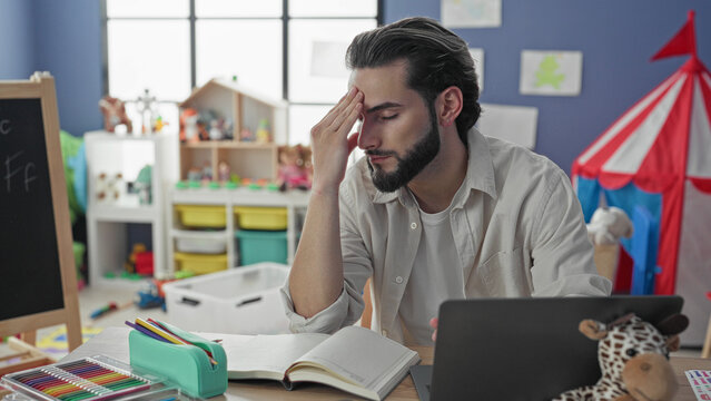 Young man teacher at desk with laptop, open book and colored pencils hand to forehead while writing notes in classroom building; teaching fatigue.