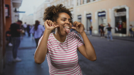 Young african american woman in a striped t shirt pulls her cheeks and sticks out her tongue while making playful faces on a crowded city street  playful expression. © Krakenimages.com