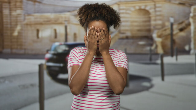 Woman sticks tongue out and frames her face with both hands while standing near bollards and a parked car on an urban street; playful mischief.