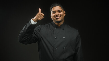 Obraz premium Hispanic man chef in black uniform showing peace sign and later thumbs up with hand in studio, smiling and wearing earring; confidence.
