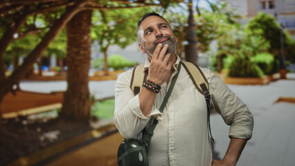 Fototapeta premium Middle age bearded man with backpack and canteen touching chin and smiling in forest walkway near park benches; playful curiosity.