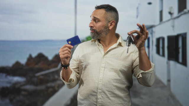 Man holds creditcard in left hand and car key in right hand by a building on the seaside walkway; indecision choices.