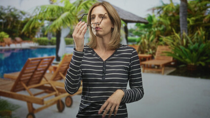 Fototapeta premium Woman pinches bridge of nose and adjusts glasses, brushing hair with fingers while wearing striped shirt in studio pool set; fatigue headache travel.